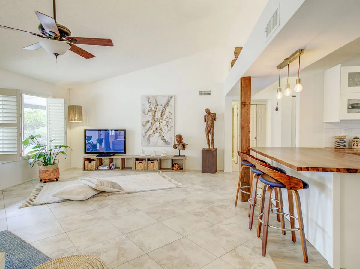 A contemporary living space featuring a television, striking artwork on the wall, a lush plant near a large window, wooden bar stools, and a unique sculpture in the corner. The room is set up for socializing, relaxing, or enjoying a movie night, with sophisticated design elements enhancing the space.