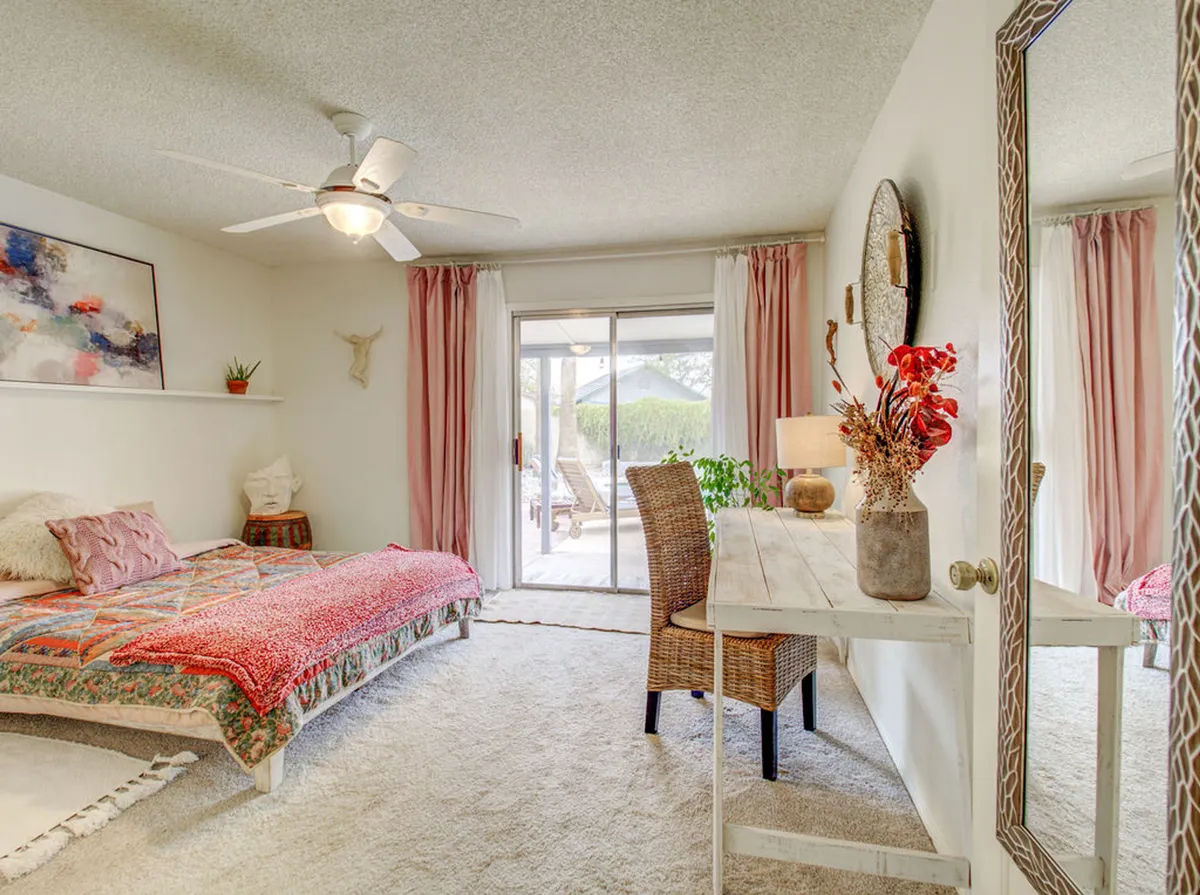 A tranquil bedroom workspace featuring a bed with a colorful quilt, a woven chair and desk beside a sliding glass door, and a decorative mirror with a flower arrangement. Sunlight streams in, creating a bright, inviting atmosphere perfect for both productivity and relaxation.