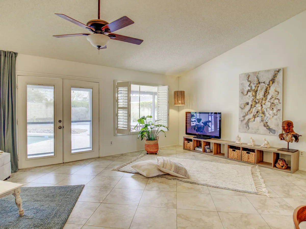 A welcoming living area featuring large windows with shutters, a comfortable TV unit decorated with stylish accents, a ceiling fan, an art piece on the wall, and a plant in a basket. Sunlight streams in, creating a bright, airy feel that highlights both relaxation and spaces designed for gathering.
