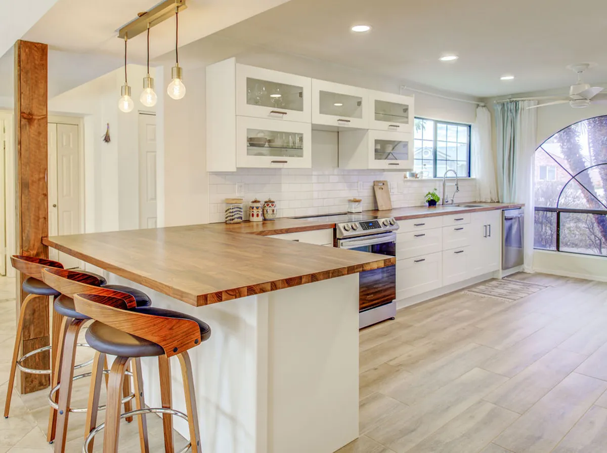 This kitchen showcases a warm, wooden island at the center, surrounded by stainless steel appliances and bathed in natural light from a large window. The pendant lights overhead and open layout create an inviting, functional space ideal for both cooking and group interaction.