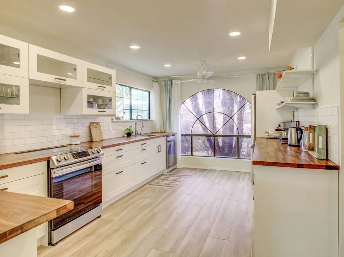 This image centers on a bright, modern kitchen featuring clean white cabinetry, expansive wooden countertops, and a large arched window letting in cheerful natural light. Modern appliances line the back wall and the peninsula-style counter offers barstool seating, inviting guests or family to gather while meals are being prepared.