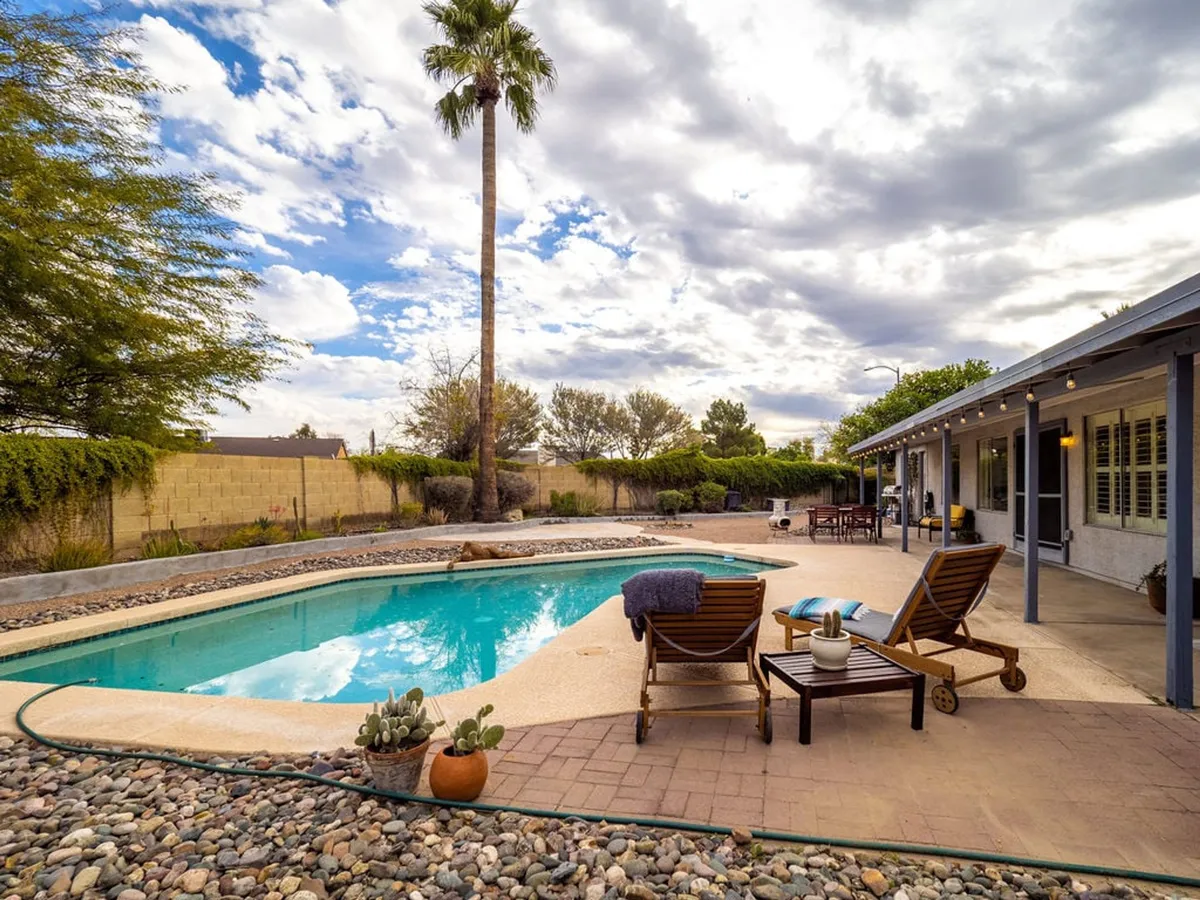 A beautifully landscaped patio area with a sparkling pool, framed by palm trees and comfortable lounge chairs. The patio space is set up for outdoor dining, and the scene suggests tranquil mornings with coffee or family swims, as well as sunset relaxation.