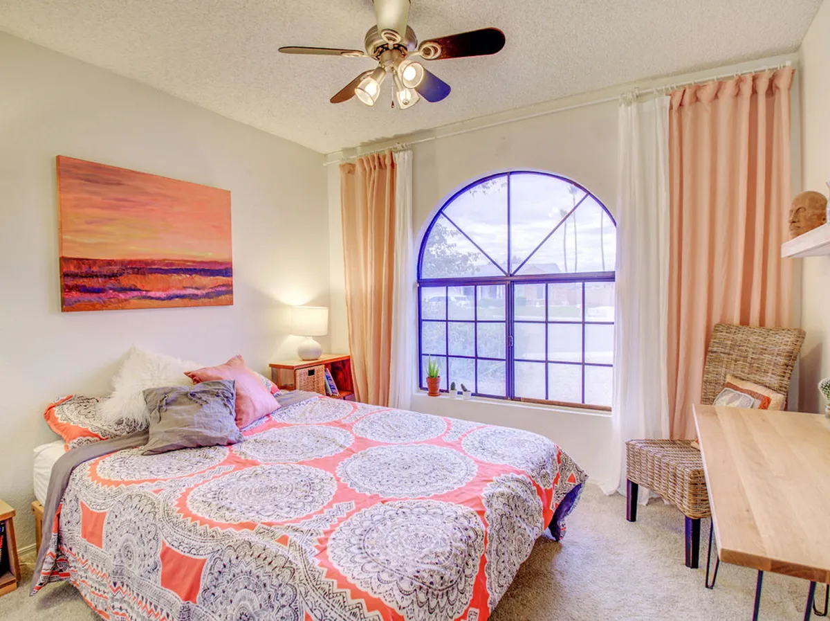 A tranquil bedroom featuring a well-made bed with colorful bedding, set beside a large arched window that allows natural light to fill the space. Subtle artwork adorns the walls, reinforcing the room’s peaceful atmosphere. The plush carpeting and soft, serene color palette enhance the sense of comfort and calmness.