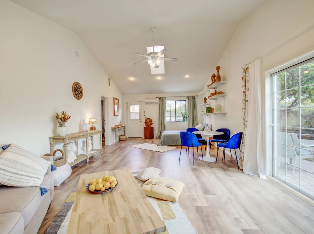 An open-concept area combining a plush sofa with decorative cushions, a round dining table surrounded by blue chairs, and a wooden coffee table with a fruit bowl. A decorative shelf with sculptures adds an artistic touch, while natural light fills the room, promoting a sense of warmth and sociability.