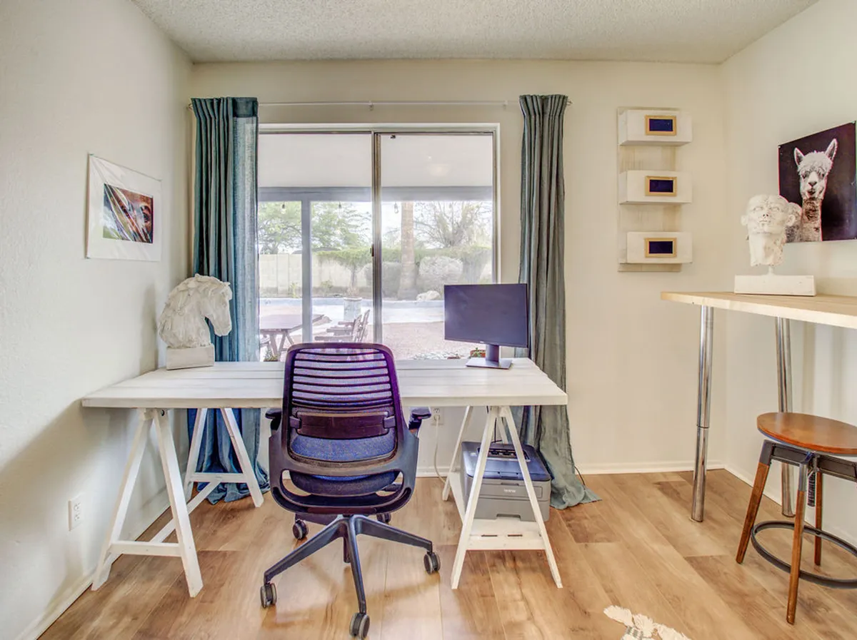 A serene and productive studio space featuring a desk with a computer monitor, window view, artwork, and decorative objects. Ample natural light streams in through the window, illuminating the work area and creating a calming environment ideal for both work and relaxation.