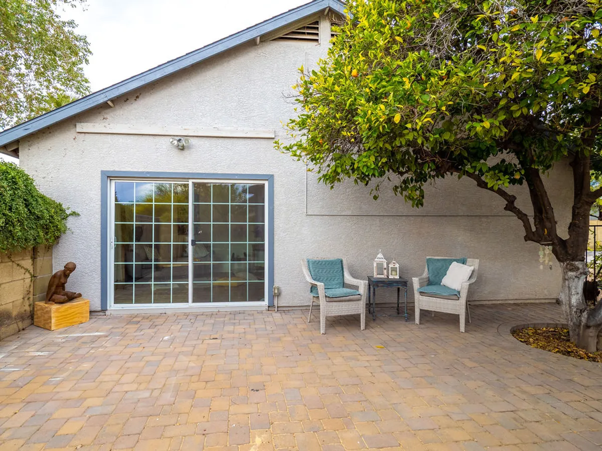 A tranquil outdoor patio area with comfortable seating nestled under a large tree, adjacent to a sliding glass door that connects the patio to the interior. Sunlight filters through the leaves, creating a peaceful spot perfect for relaxation, reading, or light conversation.