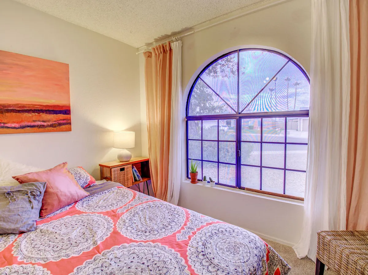 A cozy bedroom highlighted by a bed with colorful bedding, a large arched window flooding the space with natural light, and a vibrant art piece on the wall. The room feels both restful and inspiring, perfect for quiet moments or creative thinking.